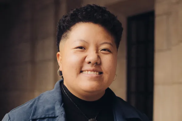 "Portrait of Chueqa Yang smiling, wearing a blue jacket, black turtleneck, and bolo tie."