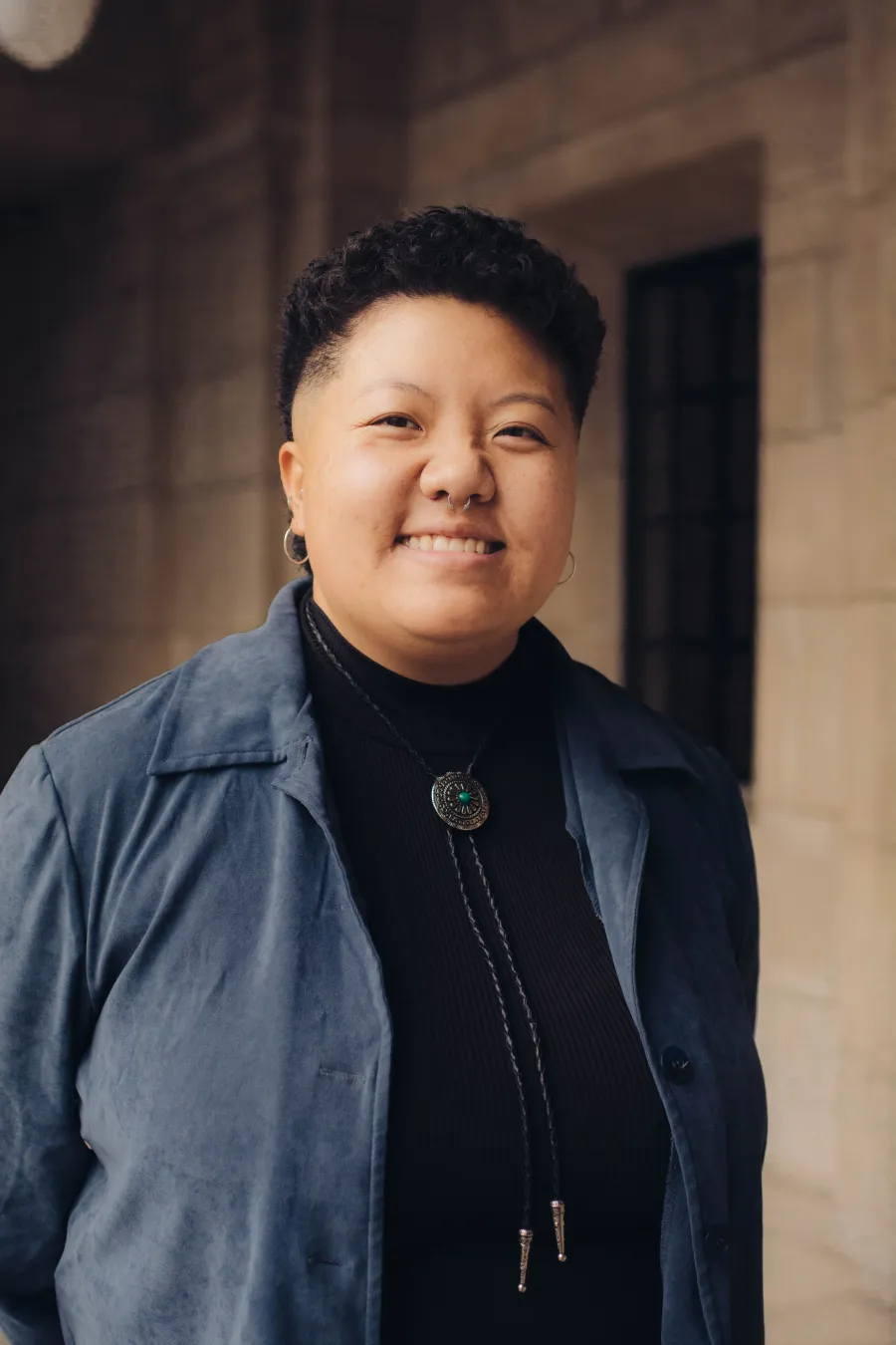"Portrait of Chueqa Yang smiling, wearing a blue jacket, black turtleneck, and bolo tie."